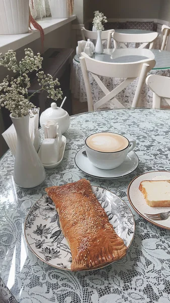 Lezzetli bir kahve dantel kaplı bir masanın üzerinde pul pul bir pasta ve bir dilim ekmeğin yanında duruyor. Kafede büyüleyici dekorasyonları ve yumuşak doğal ışığı olan rahat bir atmosfer var..
