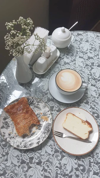 Sıcak bir fincan kahve bir dilim pasta ve dantel kaplı bir masanın üzerinde bir parça hamur işi yanında oturur. Çiçekli küçük bir vazo rahat kafe atmosferine cazibe katar..