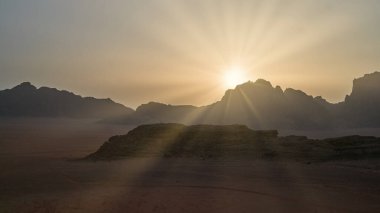 Sakin bir gündoğumu gökyüzünü engin bir çöl manzarası üzerine yumuşak ışıkla doldurur. Arka planda dağlar beliriyor, doğanın güzelliğini ve sakin atmosferini vurguluyorlar..