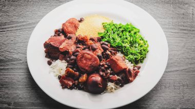 Feijoada, tipik Brezilya mutfağı. Feijoada beyaz pirinç, siyah fasulye, sosis, domuz eti, domuz pastırması, kızarmış couve ve manyok un ile beyaz bir yemek üzerinde. Ahşap masa arka plan.