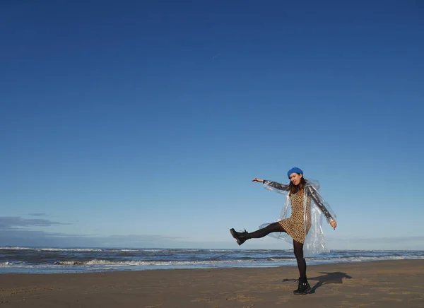 girl in the dress walks widely on the beach. Dressed in transparent raincoat, blue beret and black shoes. Day. horizontally. goes from right to left