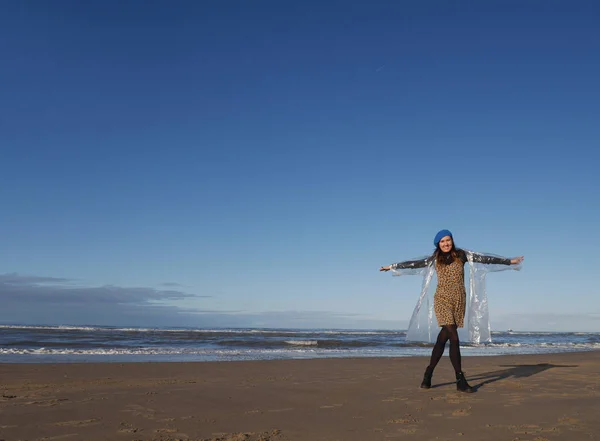 Ein Mädchen im Regenmantel steht am Strand und blickt in die Kamera. Es zeigt ein Flugzeug oder einen Vogel. blauer Himmel und Wellen. Es gibt Raum zum Schreiben. horizontal — Stockfoto