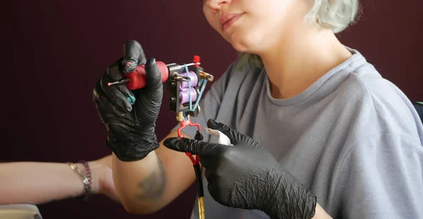 Mistrz tatuażu dziewczyna z maszyną w ręku. w czarnych rękawiczkach i szarym T-shirt. — Zdjęcie stockowe