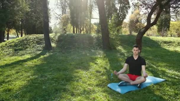 Молодой человек практикует йогу на свежем воздухе в парке, сидя на коврике в лотосном позе с закрытыми глазами, медитируя. Прорес, медленное движение — стоковое видео