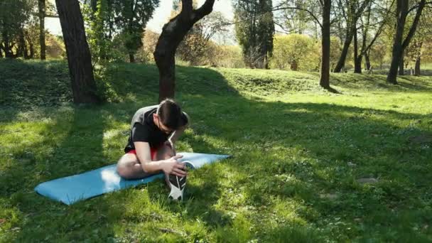 Hombre joven deportivo teniendo su yoga y estirándose temprano en la mañana en el parque soleado de verano. En una camiseta negra y pantalones cortos rojos en una alfombra azul. Prores, cámara lenta — Vídeos de Stock