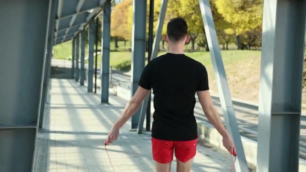 Retrato de un joven en forma con cuerda de salto en la plataforma cerca de bastidores de metal. Fitness Saltar el entrenamiento al aire libre. El tipo salta cerca de los pilares de metal en el fondo del estadio. Vestido con una T negra — Vídeo de stock