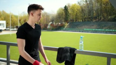 Güzel Genç Adam Kamera Görünüyor Ve İçecekler Temiz Su Susuzluk Spor tarafından Yoga ve Ateşleme Egzersizleri Performans Sonra. Kişi Yakındaki Stadyum, Ağaçların Arkasında Duruyor. Yoğun Yaz