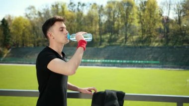 Güzel Genç Adam Kamera Görünüyor Ve İçecekler Temiz Su Susuzluk Spor tarafından Yoga ve Ateşleme Egzersizleri Performans Sonra. Kişi Yakındaki Stadyum, Ağaçların Arkasında Duruyor. Yoğun Yaz