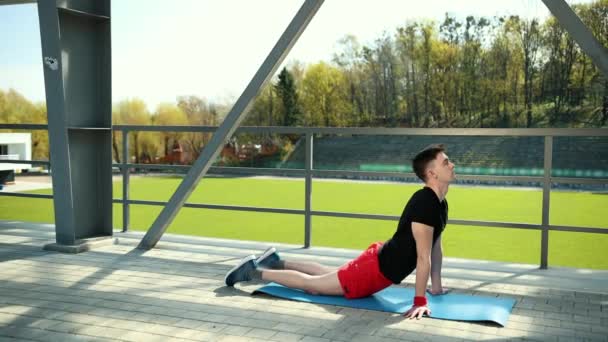 Schöner junger Mann beim Stretching der Achillessehne auf einer Matte im Stadion. — Stockvideo
