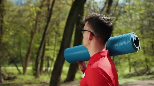 Ein gutaussehender junger Mann mit Brille und blauem Teppich für Yoga auf der Schulter sieht aus, als suche er nach einem Platz für Sport.. — Stockvideo