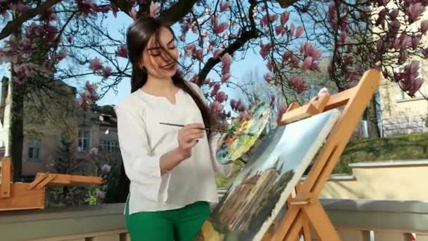 Linda artista menina pinta seu quadro no fundo de uma árvore sakura. Perto de mulheres, cavalete, ossos e tintas — Vídeo de Stock