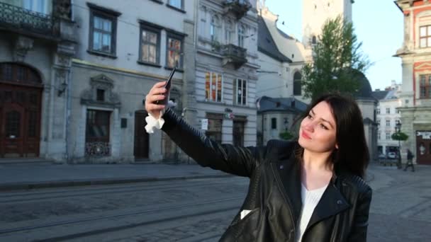 Mulher morena bonita olha para o telefone e tira selfies na cidade. Atrás de uma boa arquitetura urbana. Um blog de blogueiros leva ao vivo pelo telefone. Prores, câmera lenta — Vídeo de Stock