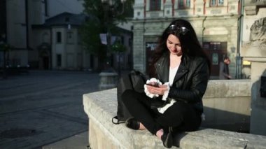 Akıllı telefonlu kuryeyle mesaj atan güzel, genç, beyaz bir kadın anıtın yanında oturuyor. Ağır çekim. Dualar
