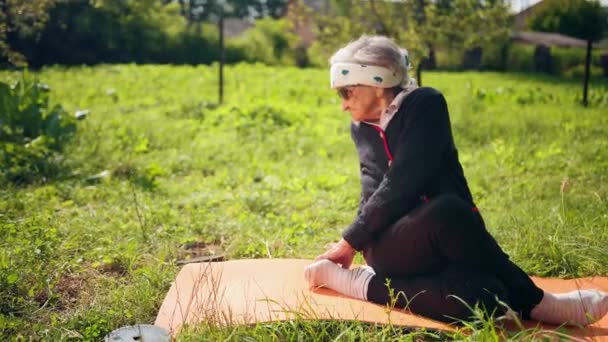 Betagte Dame auf einer Matte für Yoga stellt Fuß auf Fuß und macht Stretchgehäuse — Stockvideo