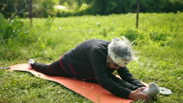 Donna molto anziana si siede su spaccature longitudinali, tiene le mani dietro la gamba e si tocca la fronte al ginocchio — Video Stock