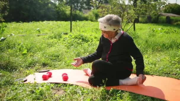 Äldre kvinna sitter på mattan i yoga pose och försöker höja Pink hantel. — Stockvideo