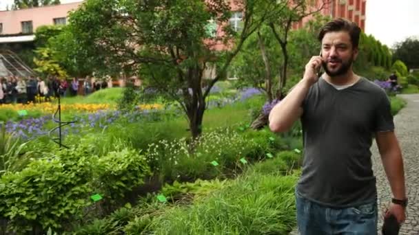 Ένας νεαρός που τηλεφωνεί κοντά σε λουλούδια στον κήπο. Φυτά στο παρασκήνιο. — Αρχείο Βίντεο