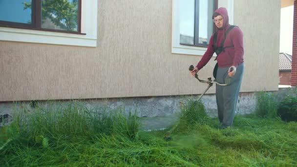 Парень садовник косит высокую зеленую траву с газонокосилкой, днем, рядом с домом — стоковое видео