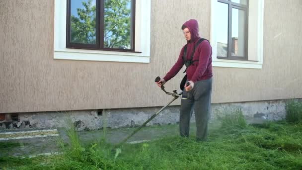Парень садовник косит высокую зеленую траву с газонокосилкой, днем, рядом с домом — стоковое видео