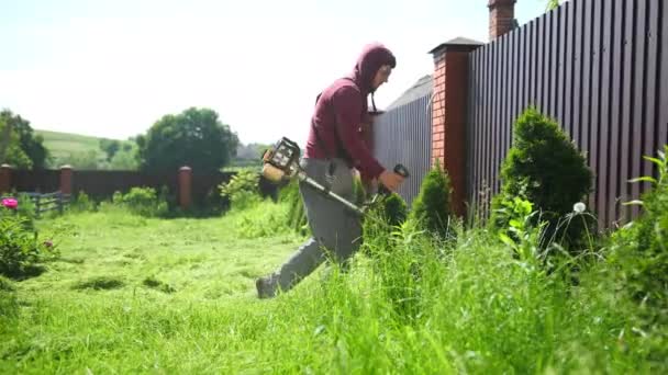 Чоловік садівник косить траву бензиновою газонокосаркою уздовж високого паркану, сонячний день — стокове відео