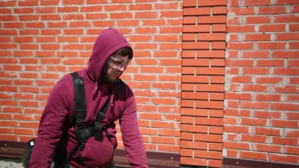 Человек в защитных очках, косит высокую газонокосилку, вдоль высокого кирпичного забора — стоковое видео