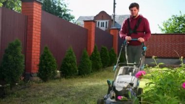 Genç adam bir çim biçme makinesi ile çim çimlerini biçiyor, yüksek bir çit boyunca, çiçekler yakın