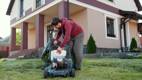 Sakallı genç bir adam, çim biçme makinesini temizliyor, büyük ve güzel bir evin yanında. — Stok video