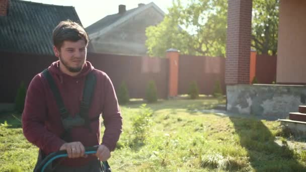 Молодой человек с бородой в солнечный день стрижет газон газонокосилкой — стоковое видео