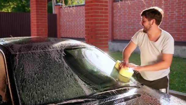 Jeune homme vêtu d'un t-shirt blanc, lave avec du savon et une voiture éponge jaune — Video