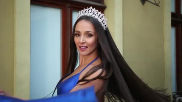 Fröken, brunett, i en blå klänning med en djup urringning, med en krona, poserar — Stockvideo