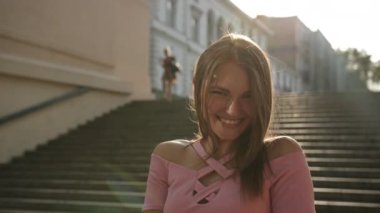 Pembe elbiseli kız, kameranın önünde gülümseyerek poz veriyor, merdivenlerde duruyor.