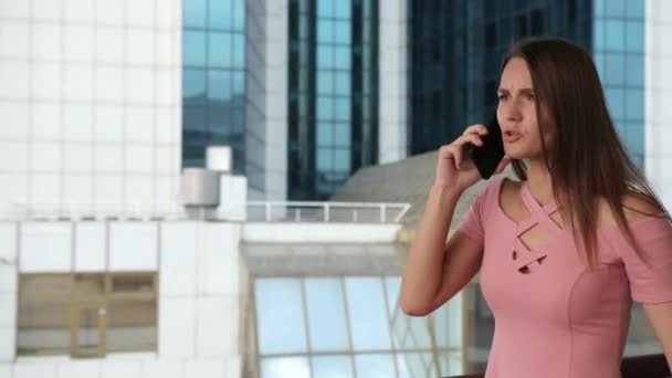 Девушка держит смартфон, в розовом платье, эмоционально разговаривает и машет руками — стоковое видео
