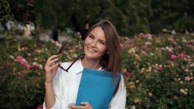 Mavi klasör tutan gözlüklü genç kız, sokakta kameraya poz veriyor.
