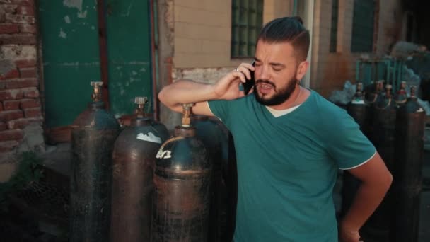 Mann hält Smartphone, steht in Fabriknähe, es gibt viele Gasflaschen — Stockvideo