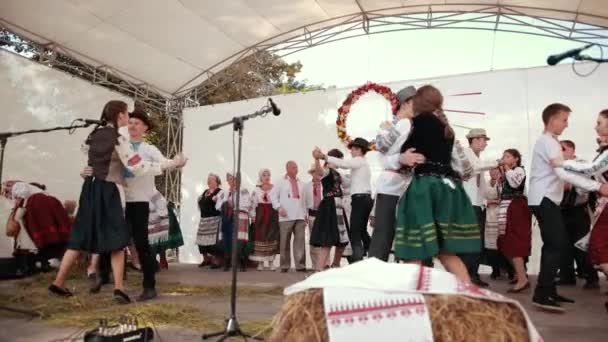 На білій сцені виступають жінки і чоловіки в народних костюмах, співають і танцюють — стокове відео