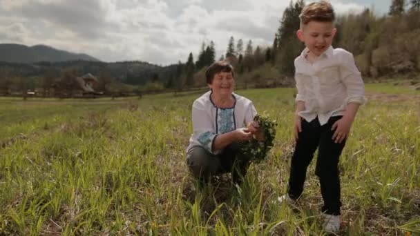 Babcia robi wieniec z dzikich kwiatów ze swoim młodym wnukiem. Spokojnie na zewnątrz. — Wideo stockowe