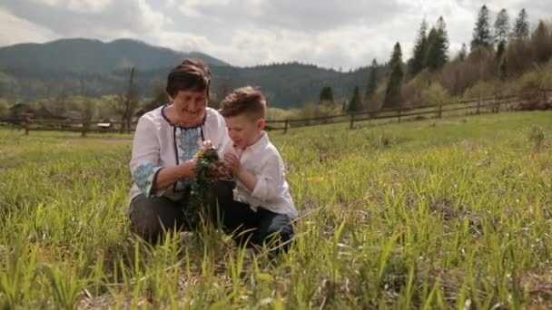 La abuela hace una corona de flores silvestres con su nieto joven. Relájate afuera. — Vídeos de Stock