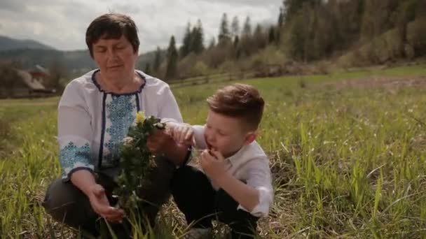 Grootmoeder maakt een krans van wilde bloemen met haar kleinzoon. Ontspan buiten. — Stockvideo