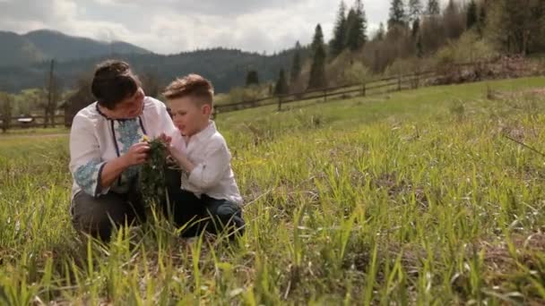 Grootmoeder maakt een krans van wilde bloemen met haar kleinzoon. Ontspan buiten. — Stockvideo