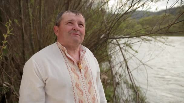 Старый пожилой человек, стоящий на берегу реки в вышитой рубашке — стоковое видео