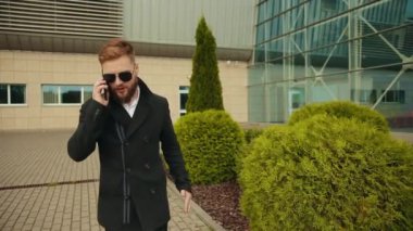 Güneş gözlüklü kızgın işadamı telefonda konuşuyor. Takım elbiseli adam yürüyor.