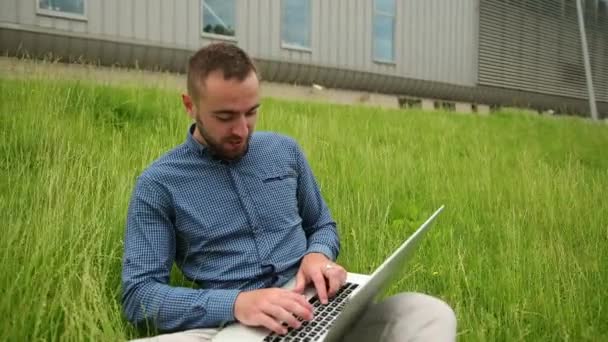 Молодой человек сидит на траве, используя ноутбук, мужчина с компьютером работает удаленно — стоковое видео