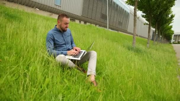 Jovem sentado na grama, usando laptop, macho com computador correndo remotamente — Vídeo de Stock