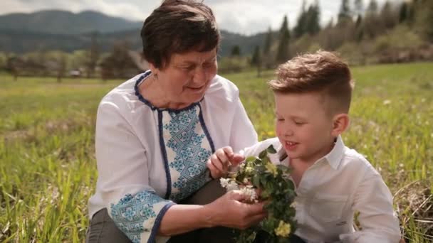 La abuela hace una corona de flores silvestres con su nieto joven. Relájate afuera. — Vídeos de Stock