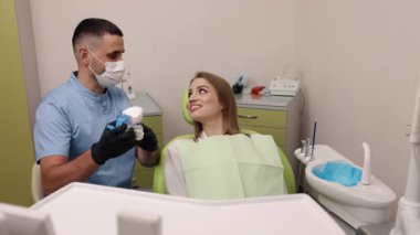 Diş implantı, ağız muayenesi, diş bakımı. Diş hekimi sandalyesindeki kadın hasta, yüz maskesi takan, diş modelini ve aletlerini tutan, tedavi prosedürünü açıklayan erkek dişçi olarak dikkatle dinliyor.