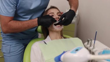 Diş muayenesi, sağlıklı dişler, diş tedavisi. Mavi önlüklü dişçi, dişçi koltuğunda ağız retraktörüyle oturan kadın dişlerini inceliyor..