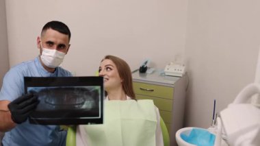 Gülümseyen hasta, diş kontrolü, dişçi sandalyesi. Diş muayenehanesinde, mavi elbiseli ve maskeli dişçi mutlu hastaya diş röntgeni gösteriyor..