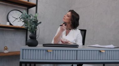 Makyaj uygulaması, kendine güvenen bir kadın, masa makyajı. Profesyonel ofis ortamında, beyaz elbiseli kadın, iş yerinde yüzüne makyaj fırçası sürüyor..