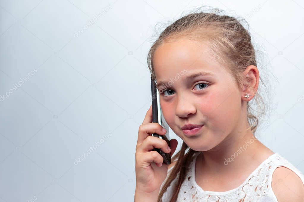 A portrait of a pretty young caucasian girl (10 years old) wearing a white summer dress or blouse making a phone call using a cellphone looking at the camera on a light background