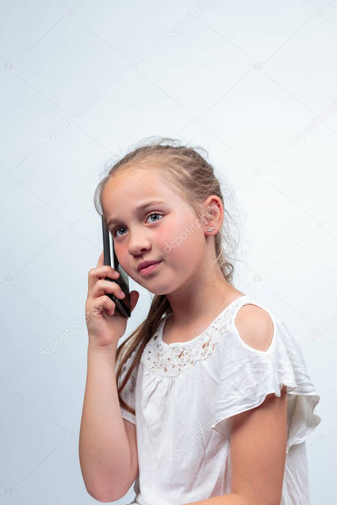 A portrait of a pretty young caucasian girl (10 years old) wearing a white summer dress or blouse making a phone call using a cellphone looking away from camera on a light background